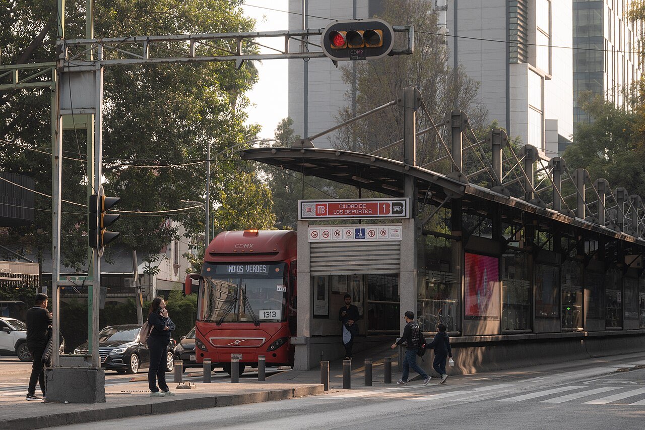 A red Metrobús bus pulling up to the Ciudad de los Deportes station on a Mexico City street