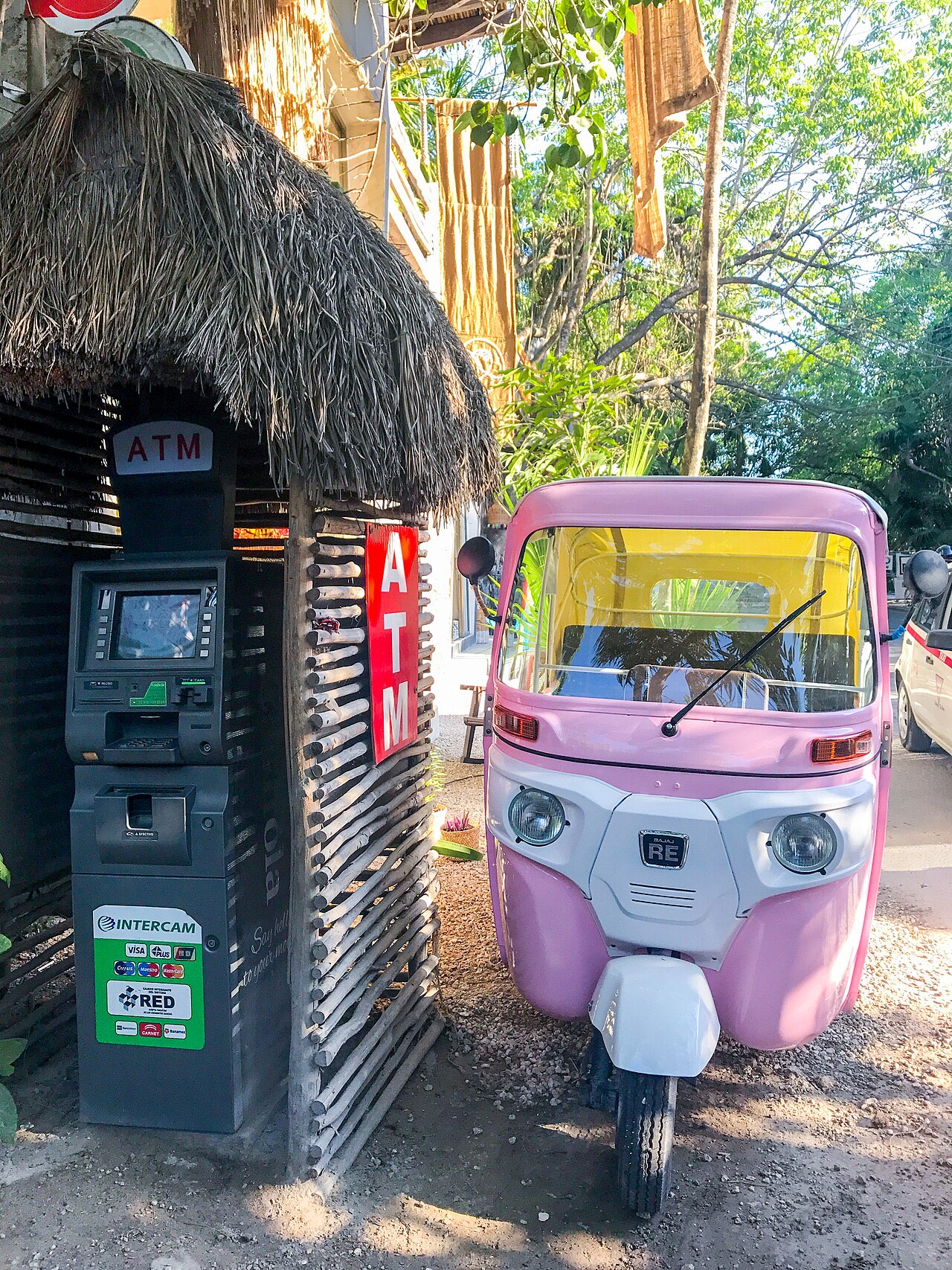 An ATM under a thatched shelter in Tulum with card-network logos on the machine, beside a pink auto-rickshaw