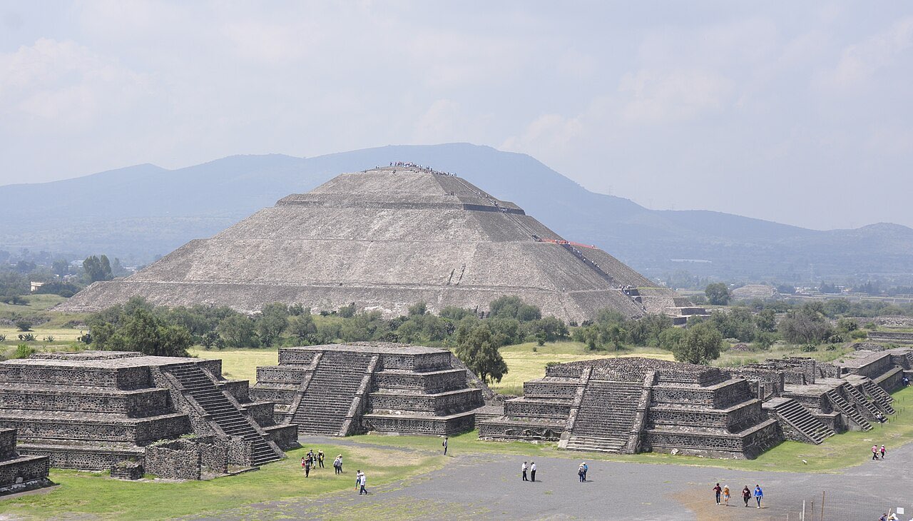 Stepped temple platforms in the foreground with the broad Pyramid of the Sun behind them under a hazy sky